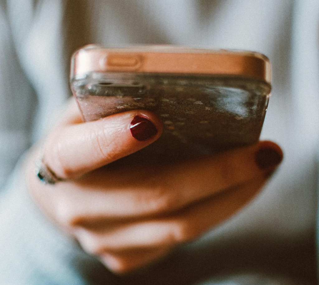 Närbild på en person som håller en mobiltelefon med rosa skal i händerna. Personen har mörkrött nagellack.