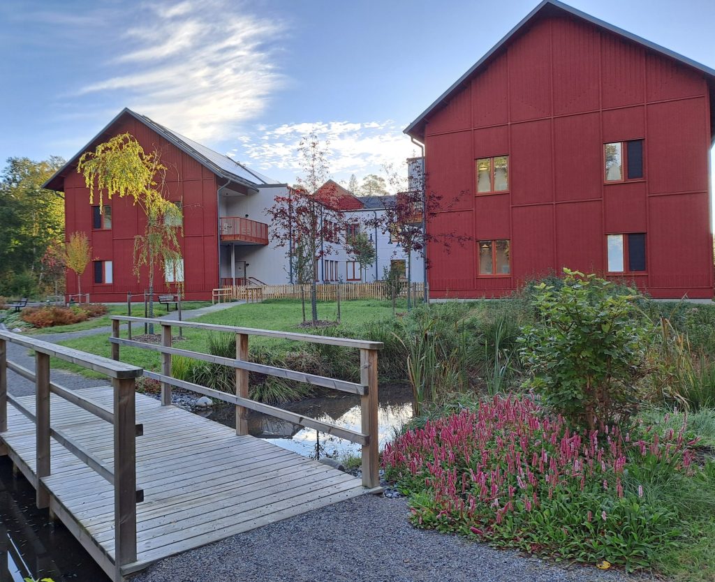 Röda hus med en gångbro i förgrunden, föreställer äldreboendet Pilhamns gårdar