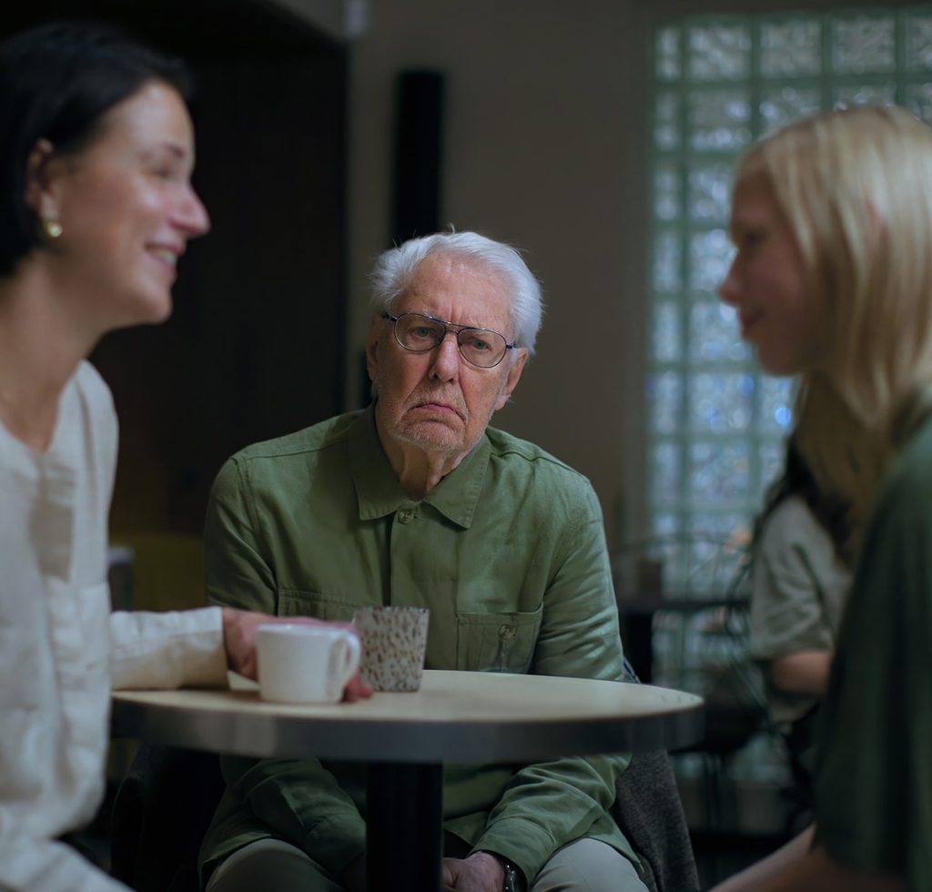 Foto på en man som ser ensam ut i sällskap med två personer som ler mot varandra. Fotot visar existentiell ensamhet i syfte att gestalta projektet Tankar om natten.